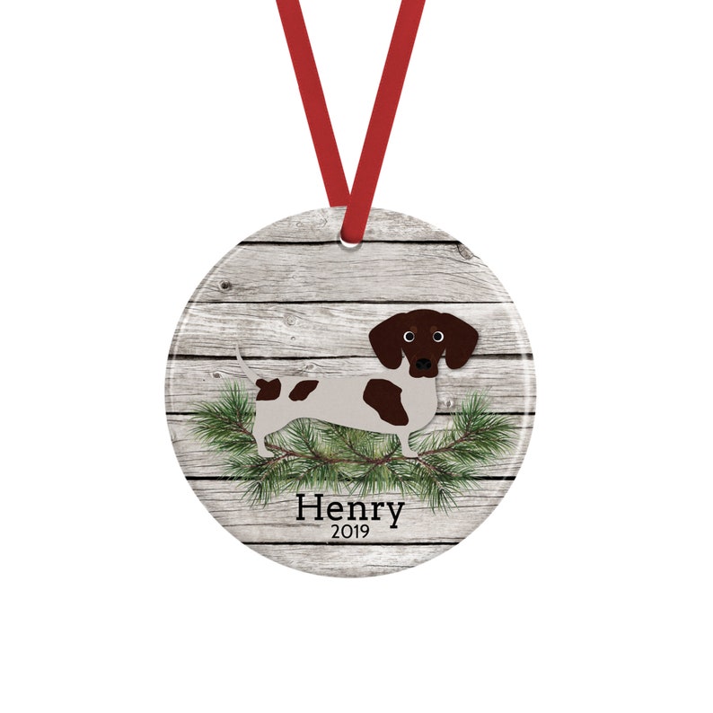 May include: A round ceramic ornament featuring a brown and white dachshund illustration over a gray wood grain background with green pine needles. The text Henry 2019 is printed in black below the dog. A red ribbon is attached for hanging.