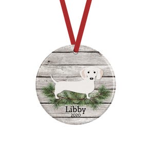 May include: A round ceramic ornament featuring a white dachshund illustration over a gray wood grain background with green pine needles. The text Libby 2020 is printed in black below the dog. A red ribbon is attached to the top for hanging.
