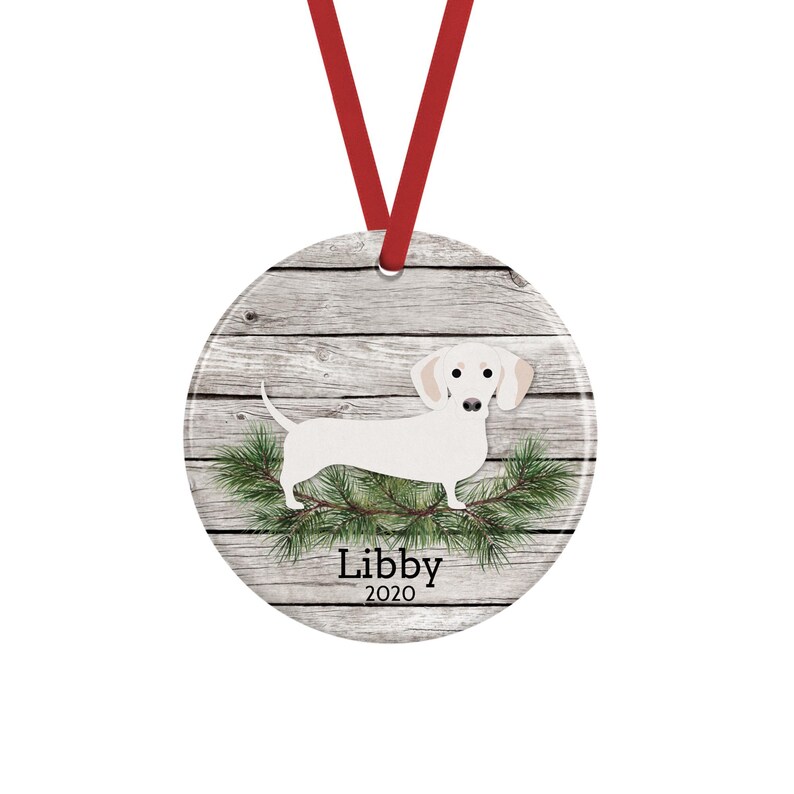 May include: A round ceramic ornament featuring a white dachshund illustration over a gray wood grain background with green pine needles. The text Libby 2020 is printed in black below the dog. A red ribbon is attached to the top for hanging.