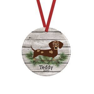 May include: A round ceramic ornament featuring a brown and tan dachshund standing on green pine needles against a light gray wood grain background. The text Teddy 2020 is printed in black below the dog. A red ribbon is attached to the top.
