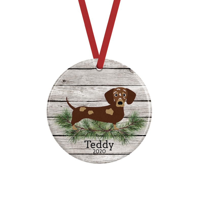 May include: A round ceramic ornament featuring a brown and tan dachshund standing on green pine needles against a light gray wood grain background. The text Teddy 2020 is printed in black below the dog. A red ribbon is attached to the top.