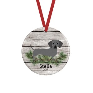 May include: A round ceramic ornament featuring a gray dachshund illustration standing on green pine branches against a light gray wood grain background. The name Stella and the year 2019 are printed in black text. A red ribbon is attached to the top.