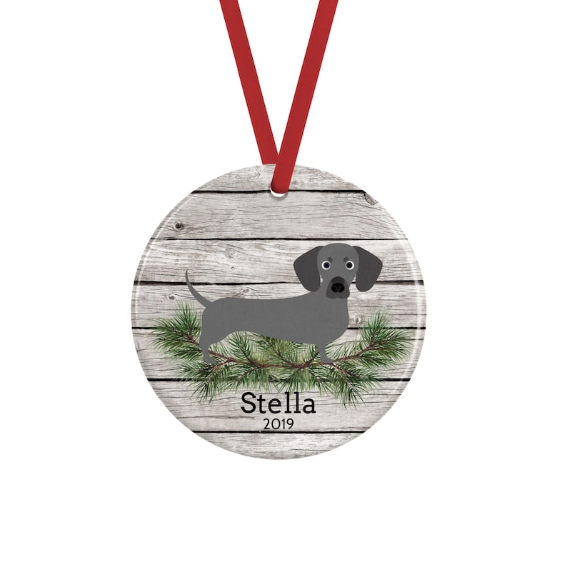 May include: A round ceramic ornament featuring a gray dachshund illustration standing on green pine branches against a light gray wood grain background. The name Stella and the year 2019 are printed in black text. A red ribbon is attached to the top.