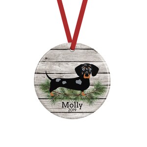 May include: A round ceramic ornament featuring a black and tan dapple dachshund illustration over a rustic gray wood plank background with green pine needles. The name Molly and the year 2019 are printed in black text. A red ribbon is attached for hanging.
