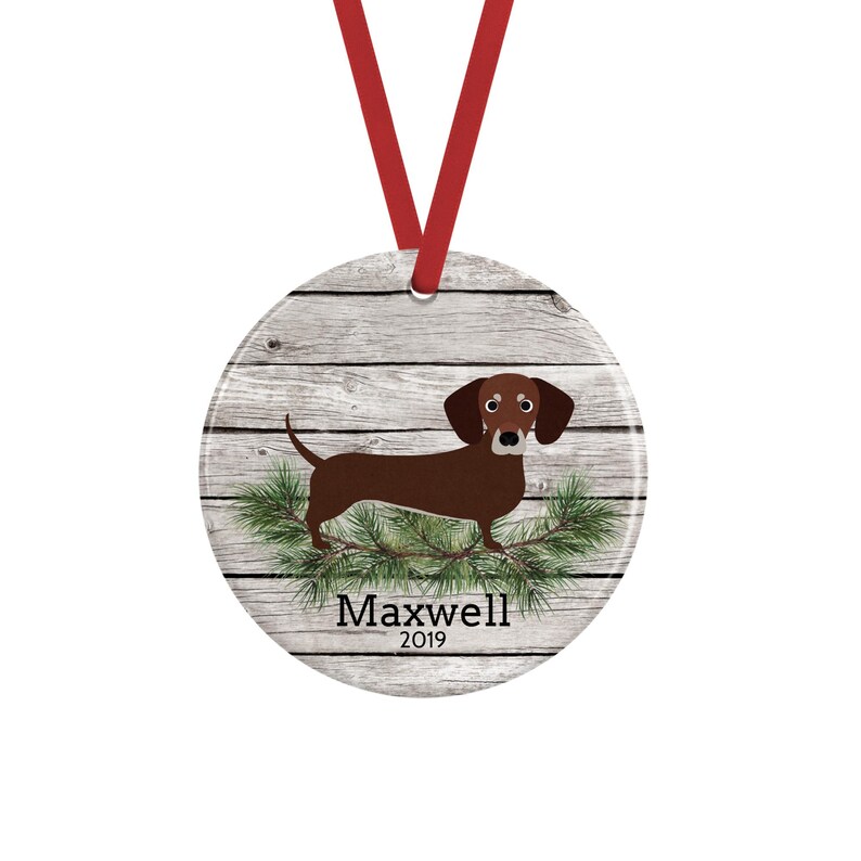 May include: A round ceramic ornament featuring a brown dachshund standing on green pine branches against a light gray wood grain background. The ornament includes a red ribbon for hanging and the text Maxwell 2019 printed in black.