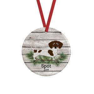 May include: A round ceramic ornament hanging from a red ribbon, featuring a white and brown spotted dachshund standing on green pine needles against a gray wood-plank background. The text Spot and 2019 are printed in black below the dog.