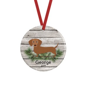 May include: A round ceramic ornament featuring a brown dachshund illustration over a gray wood grain background with green pine needles. The text George and 2019 are printed in black below the dog. A red ribbon is attached for hanging.