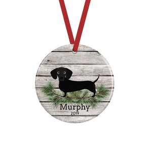 May include: A round ceramic ornament featuring a black dachshund illustration over a gray wood grain background. Green pine needles are positioned below the dog. The text "Murphy" and "2019" are printed in black. A red ribbon is attached to the top.