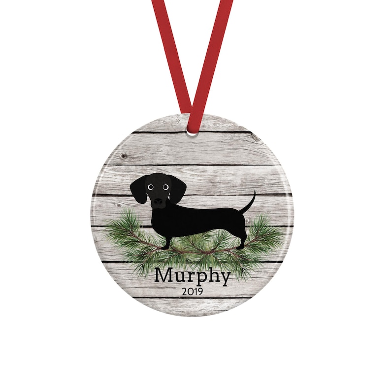 May include: A round ceramic ornament featuring a black dachshund illustration over a gray wood grain background. Green pine needles are positioned below the dog. The text "Murphy" and "2019" are printed in black. A red ribbon is attached to the top.