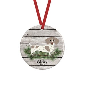 May include: A round ceramic ornament hanging from a red ribbon, featuring a white and brown spotted dachshund illustration over a gray wood grain background, green pine needles, and the text Abby 2019 printed in black at the bottom center.