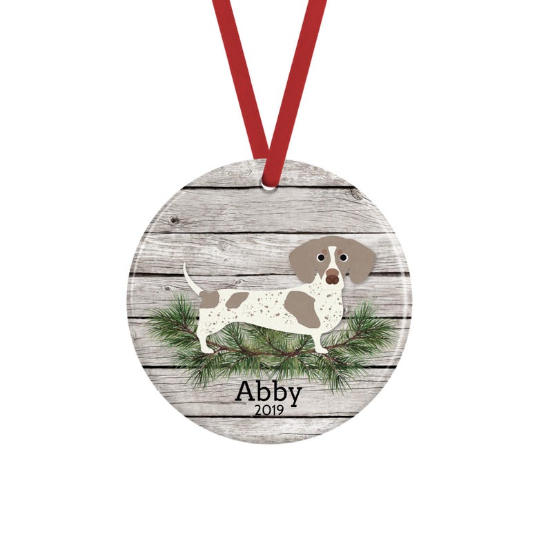 May include: A round ceramic ornament hanging from a red ribbon, featuring a white and brown spotted dachshund illustration over a gray wood grain background, green pine needles, and the text Abby 2019 printed in black at the bottom center.