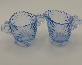 May include: A set of two blue glass creamer and sugar bowls with scalloped edges and three feet. The bowls have a delicate, vintage look.