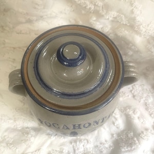 May include: A light gray stoneware crock with a lid, featuring two side handles and a brown rim. The word POCAHONTAS is printed in blue capital letters on the front. The ceramic surface has a speckled texture with blue circular accents.