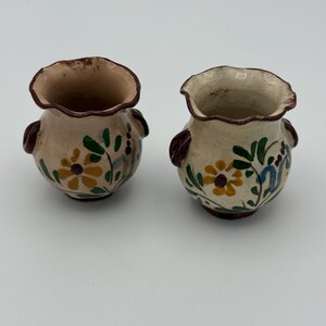May include: Two small, white ceramic pots with brown rims and painted floral designs in yellow, green, and blue. The pots have small brown handles on each side.