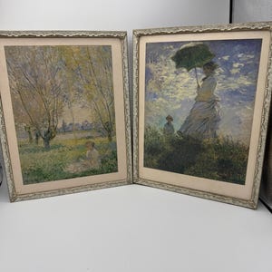 May include: Two framed paintings with silver frames. The painting on the left depicts a landscape with trees and a river. The painting on the right depicts a woman with an umbrella in a field.