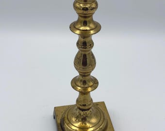 May include: A gold metal candlestick with a square base and a tall, slender stem. The candlestick has a simple, elegant design.