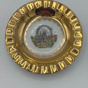 May include: A gold-rimmed porcelain ashtray with a colorful image of the Wiener Prater Ferris wheel in Vienna, Austria. The ashtray has a scalloped edge and a white center with gold accents.