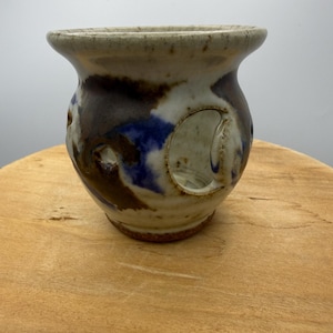 May include: A handmade ceramic candle holder with a wide, flared rim. The pottery features a blend of brown, blue, and white glaze with cut-out designs. The candle holder sits on a wooden surface.