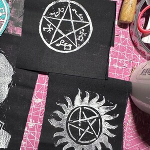 May include: Two black fabric patches with white embroidered designs. The first patch features a five-pointed star with symbols inside the points. The second patch features a sun with a five-pointed star inside.