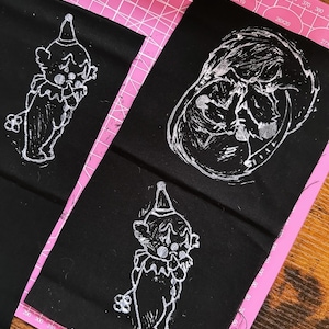 May include: Three black fabric squares with white designs. The designs are of a clown, a skull, and another clown. The fabric squares are laid out on a pink cutting mat.