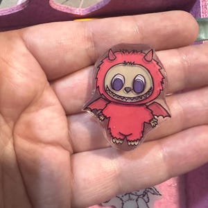 May include: A red, cartoonish monster sticker with a smiling face, horns, and bat-like wings. The sticker is held in a hand, with a pink background and other small stickers visible.
