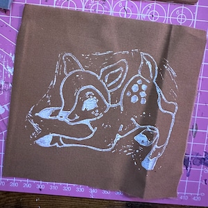 Puede incluir: Un contorno blanco de un cervatillo acostado sobre un fondo de tela marrón.