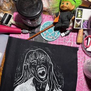 May include: A black fabric with a white linocut print of a woman screaming. The woman has long hair and is wearing a necklace. The fabric is on a pink cutting mat.