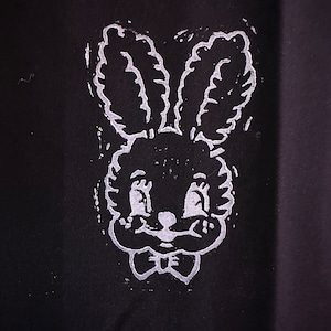 Pode incluir: Um contorno branco de um coelho com orelhas grandes e uma gravata borboleta em um fundo preto.