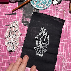 Puede incluir: Una tela negra con un diseño de troll blanco impreso en ella. La tela está sobre una estera de corte rosa con un sello de troll blanco al lado.