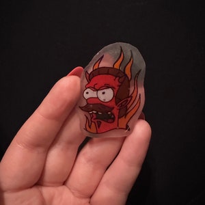 Peut inclure: Une épingle en résine transparente représentant un personnage de dessin animé à la peau rouge, des cornes et une moustache. Le personnage est entouré de flammes et a une expression de colère.