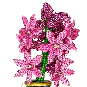 May include: A pink beaded hyacinth flower arrangement in a brown and blue ceramic pot. The flower is made of small pink beads and has a green stem.