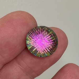 May include: A round, iridescent glass cabochon with a pink, green, and blue color scheme. The cabochon has a starburst pattern.