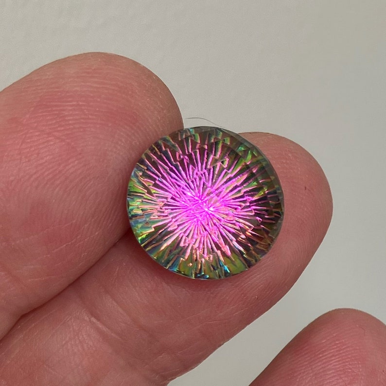 May include: A round, iridescent glass cabochon with a pink, green, and blue color scheme. The cabochon has a starburst pattern.