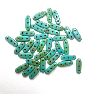May include: A collection of turquoise and green oval-shaped beads with three holes each. The beads have a mottled, aged appearance, and are scattered on a white surface. These beads are suitable for jewelry making and crafting.