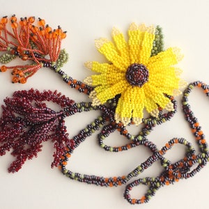 May include: A beaded necklace featuring a sunflower pendant and two smaller beaded flower accents. The necklace is made with black, orange, green, and maroon beads.