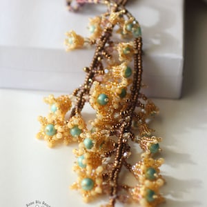 May include: A long beaded necklace with a cascading design of small flower-shaped beads in shades of gold, brown, and teal. The necklace is called "Valley of the Flowers Lariat" and is made by Rose Blu Beads.
