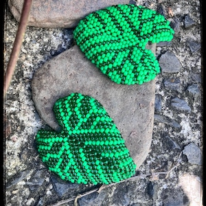 May include: Three beaded leaf-shaped earrings in bright green and dark green, resting on a gray stone. The earrings have a detailed pattern and are displayed outdoors.