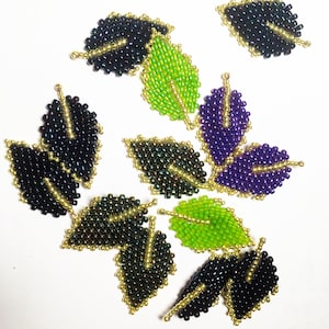 May include: A collection of eight beaded leaf shapes in various colors, including green, purple, and black. The leaves are made with small beads and have a metallic sheen.