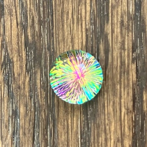 May include: A round, iridescent glass cabochon with a colorful, starburst pattern. The cabochon is set against a wood background.