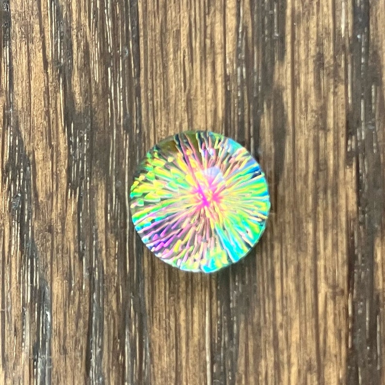 May include: A round, iridescent glass cabochon with a colorful, starburst pattern. The cabochon is set against a wood background.