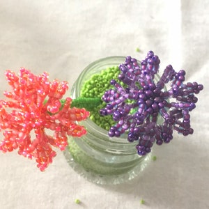May include: Two beaded flower stems in a glass jar. The flowers are pink and purple and are made of small beads. The jar is filled with green beads.