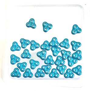 May include: A collection of small, turquoise-colored, three-lobed beads with three holes in each lobe. The beads are scattered inside a clear, rectangular container, creating a visually appealing arrangement. The beads have a metallic sheen.
