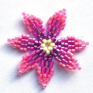 May include: A pink and purple beaded flower with a yellow centre. The flower is made with small, square beads and has six petals.