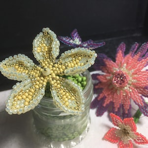 May include: Close-up of three beaded flowers. The largest flower is pink and purple, the middle flower is yellow and white, and the smallest flower is red and white. All three flowers are made with small beads and are sitting on a table.