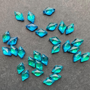 May include: A collection of small, faceted, teal and blue glass gems scattered on a dark surface. The gems have a diamond-like shape with a mix of teal and blue hues, creating a sparkling effect. Ideal for crafting or jewelry making.