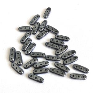 May include: A collection of dark gray, oblong beads with three holes each, scattered on a white surface. The beads have a metallic sheen and a slightly rounded shape, ideal for jewelry making or crafting projects.