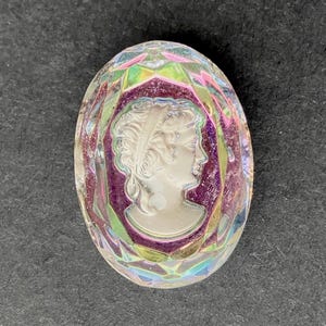 May include: Oval-shaped glass cameo with a profile of a woman. The cameo is white and set against a purple background. The glass has a faceted, iridescent finish with pink, green, and blue hues.