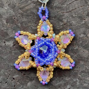May include: A beaded pendant in a starburst design. The pendant features a central blue gem surrounded by blue beads. The starburst points are adorned with gold beads and iridescent pink and purple gems. A silver loop is at the top for hanging.