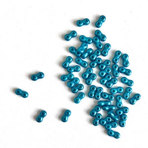 May include: A collection of teal-colored, double-holed beads scattered on a white surface. Each bead has a figure-eight shape, with two holes on each end. The beads are made of a shiny material, reflecting light. Ideal for jewelry making and crafts.
