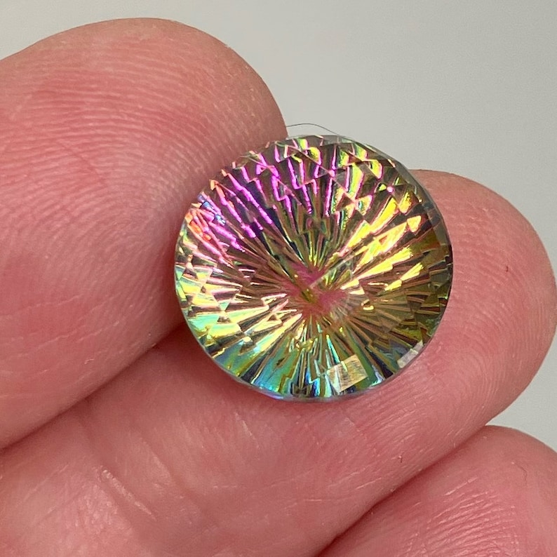 May include: A round, iridescent gemstone with a faceted, starburst design. The gemstone is clear with a rainbow of colors, including pink, yellow, green, and blue.